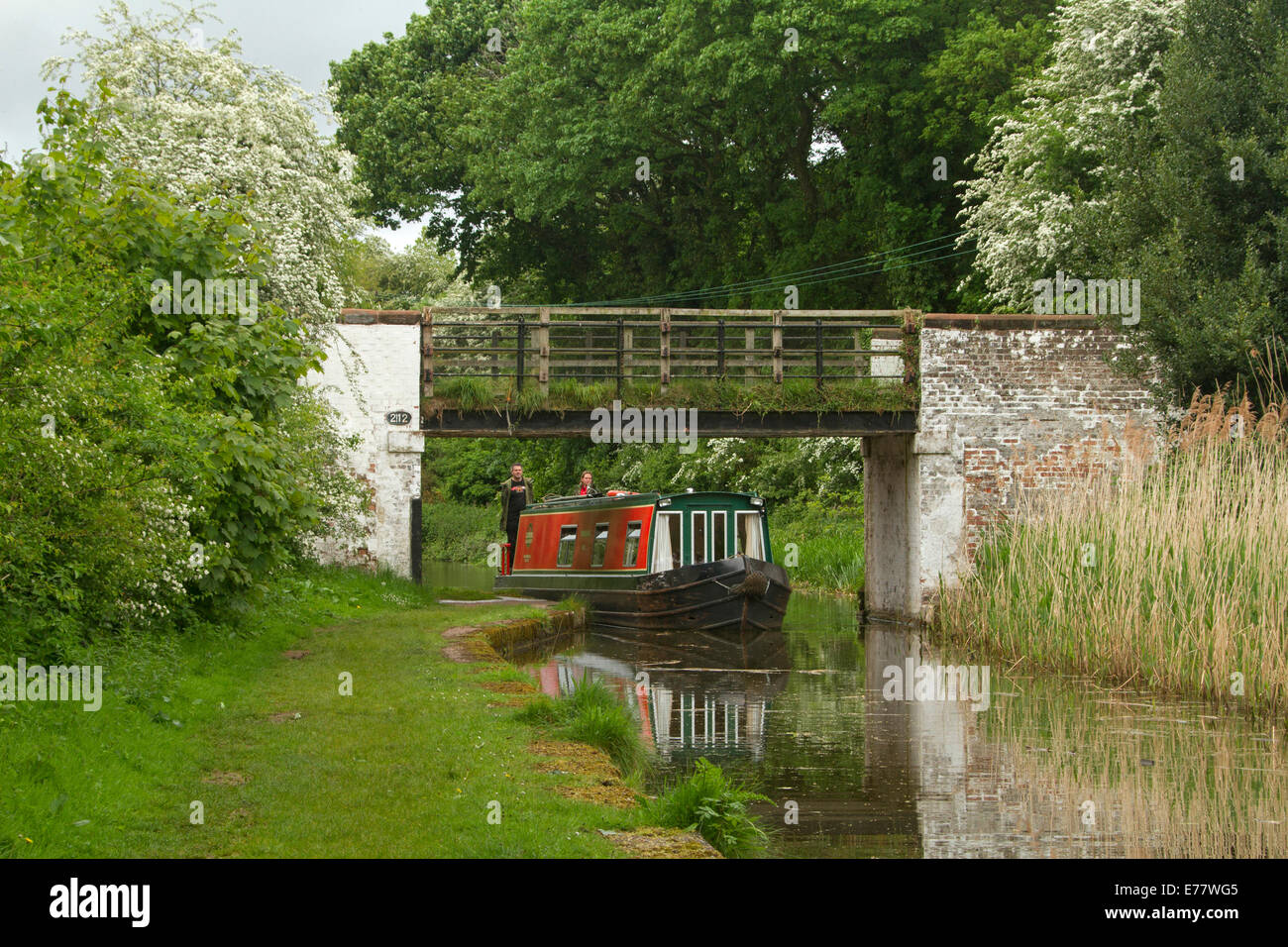 Traditional narrowboat on canal passing woodlands and under old bridge with tranquil scene reflected in mirror surface of water Stock Photo