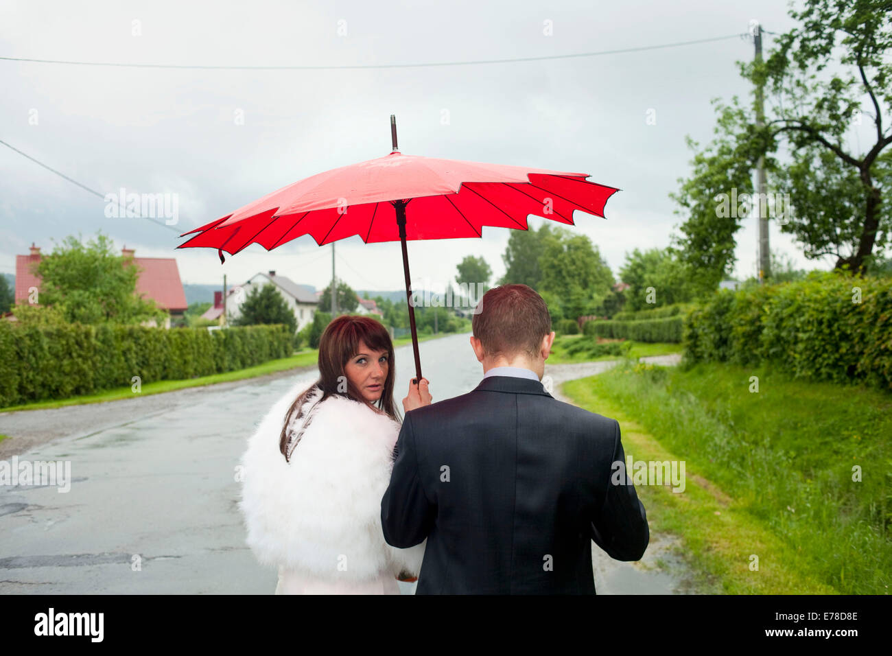 husband and wife walk down a rainy road to a formal occasion while the wife holds up a red umbrella to protect them both. Stock Photo