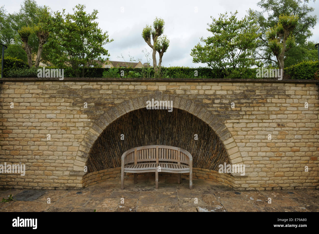 A curved wooden bench next to a Cotswold stone wall with a semi circular alcove in Rural Gloucestershire, England,UK Stock Photo