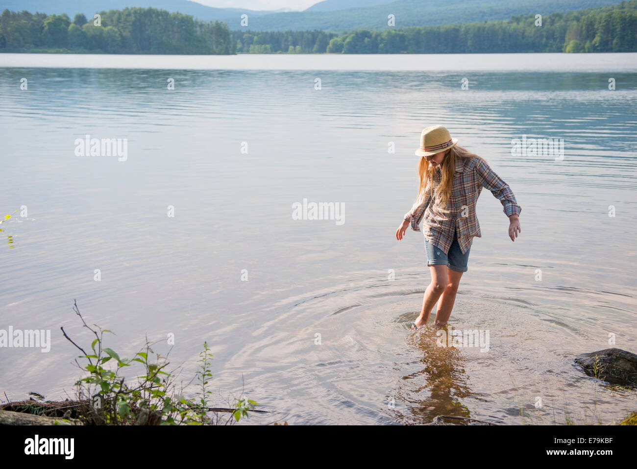 A young girl in a straw hat and shorts paddling in the shallow waters of a lake. Stock Photo