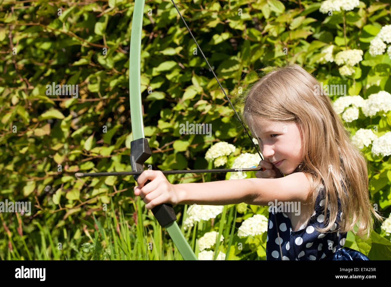 little caucasian girl aiming arrow from big bow closeup on outdoor nature background Stock Photo