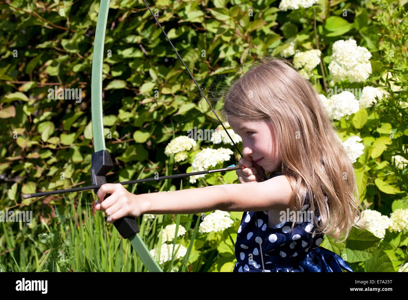 little caucasian girl aiming arrow from big bow on green garden outdoor background Stock Photo