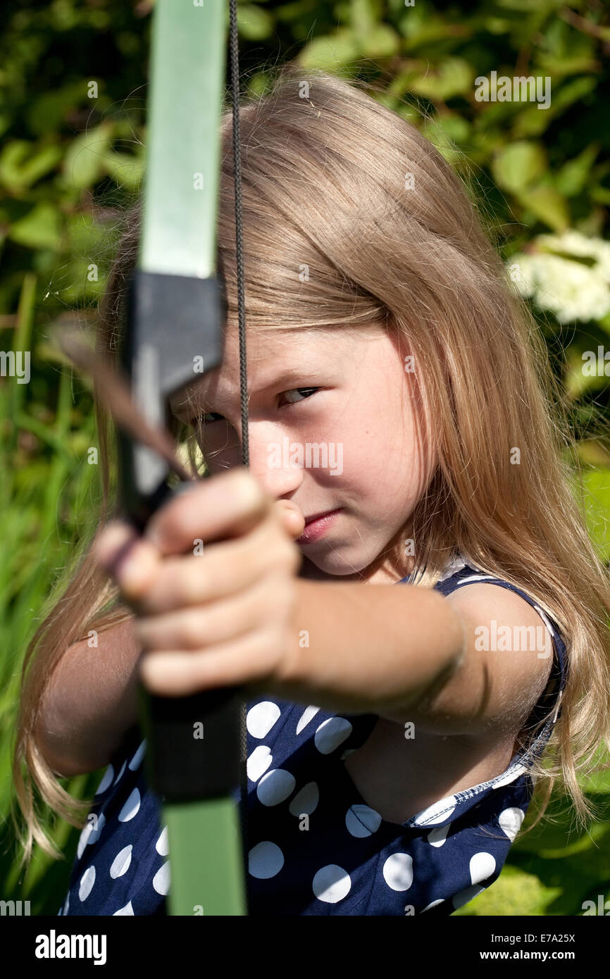 little caucasian girl aiming arrow from big bow on green garden outdoor background Stock Photo
