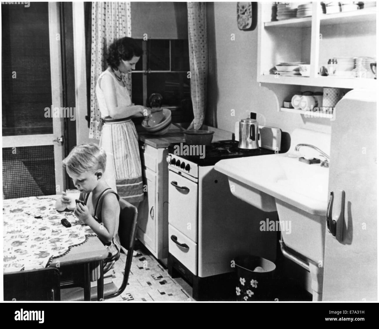 Woman Cooking in Kitchen with Young Boy Eating Cookies with Milk, USA, circa 1955, from the Documentary Film, 'Emerging Woman', Women's Film Project, 1974 Stock Photo
