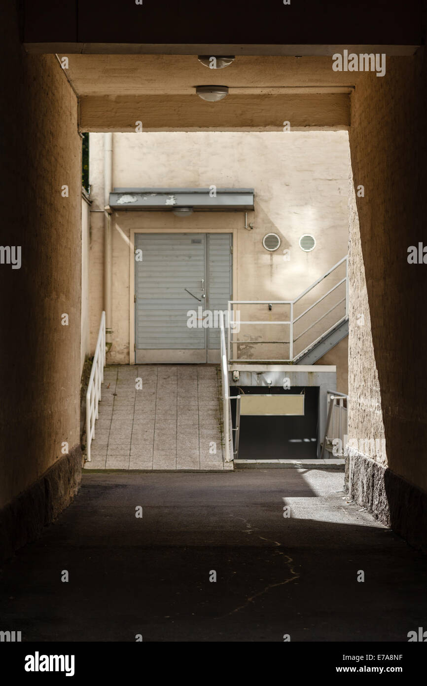 Looking through gateway in an old building towards empty alley. Concrete and cold stone walls in sunlight. Stock Photo