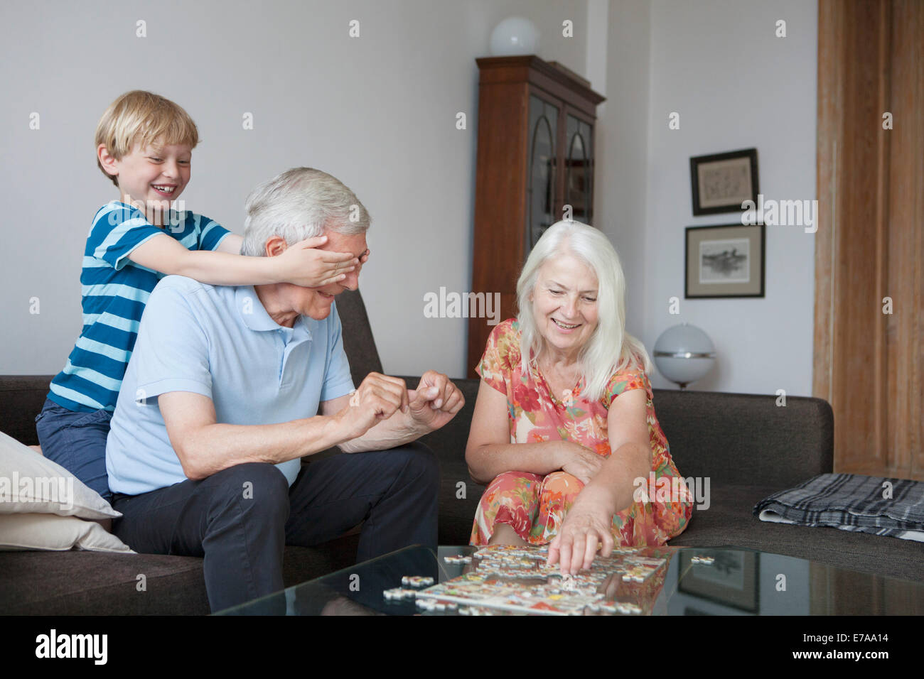Boy covering grandfather's eyes while senior woman arranging jigsaw puzzle at table in living room Stock Photo