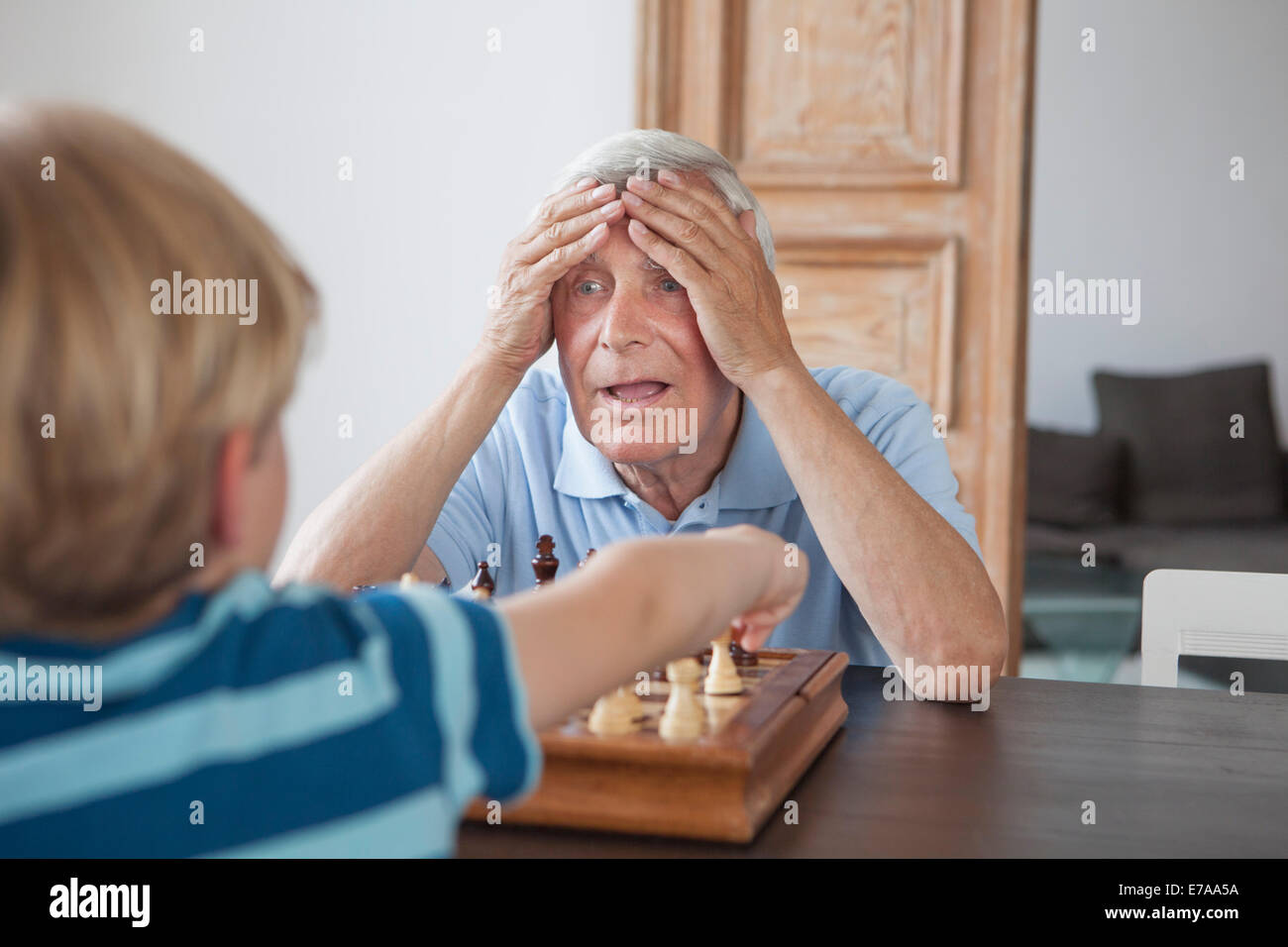 Shocked senior man with head in hands looking at grandson while playing chess at home Stock Photo