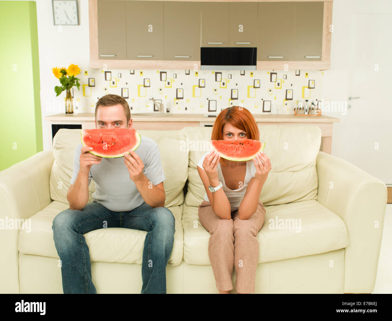 two young caucasian people sitting on sofa, holding slice of watermelon in front of their faces Stock Photo