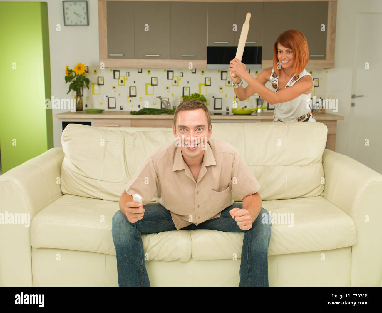 young caucasian excited man sitting on couch watching television, with woman in background trying to hit him with wooden rolling Stock Photo