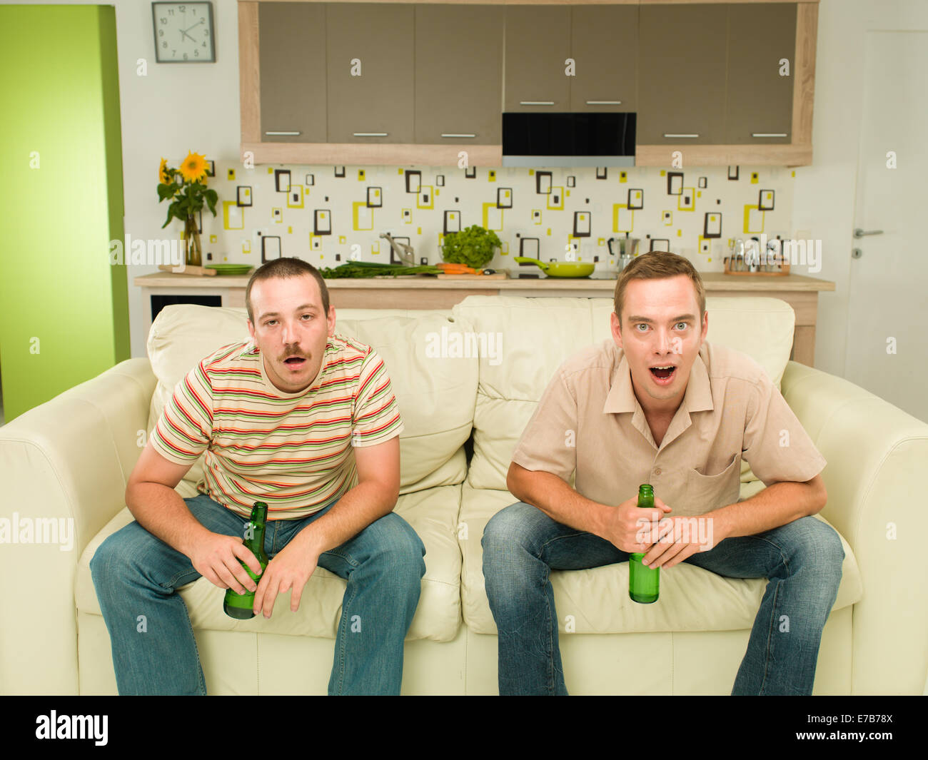 two caucasian men sitting on couch watching television and holding beers, in kitchen, one of them acting surprised, the other dr Stock Photo