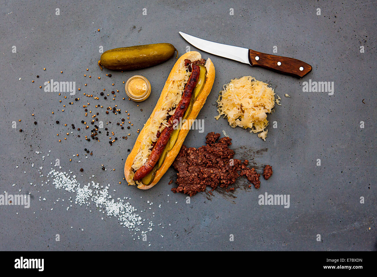 Modern hot dog with lamb sausage, sauerkraut, pickled cucumber, mustard, black sausage, salt, pepper on concrete table Stock Photo