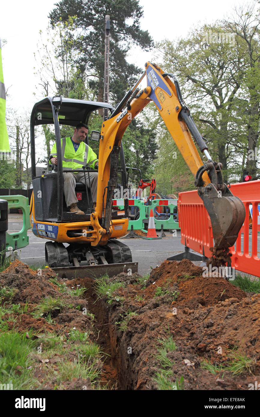 A small excavator digs a trench for new fibre optic cables next to a rural road in southern England Stock Photo
