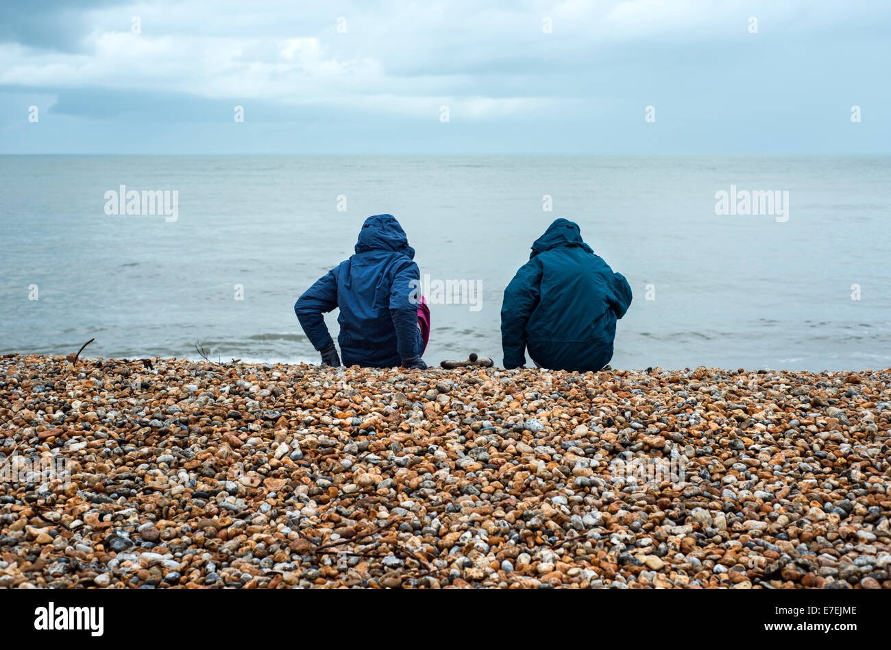 Two people wearing raincoats sat on the pebble beach and looking at the sea at Lyme Regis, Dorset, England, UK Stock Photo