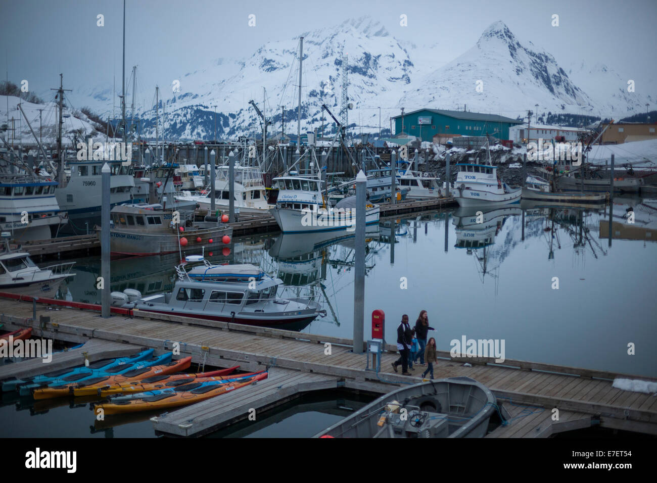 A family walks along a pier amongst sea kayaks and fishing boats at the harbor in Valdez, Alaska. Peaks of the Chugach Mountains are visible across the fjord. Stock Photo