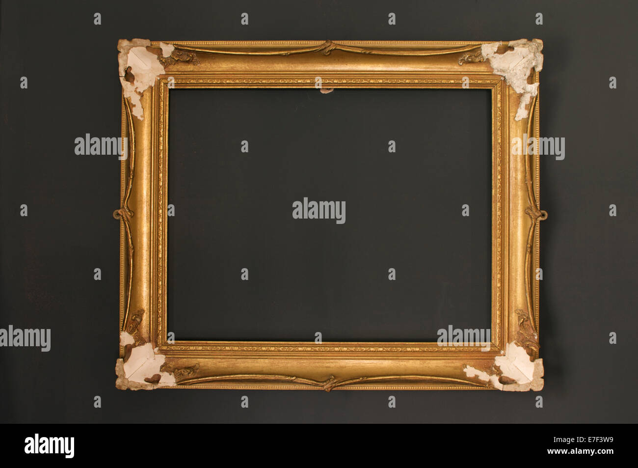 An empty distressed gilded plaster frame hanging on a grey wall which has elements of the orange paint underneath showing. Stock Photo