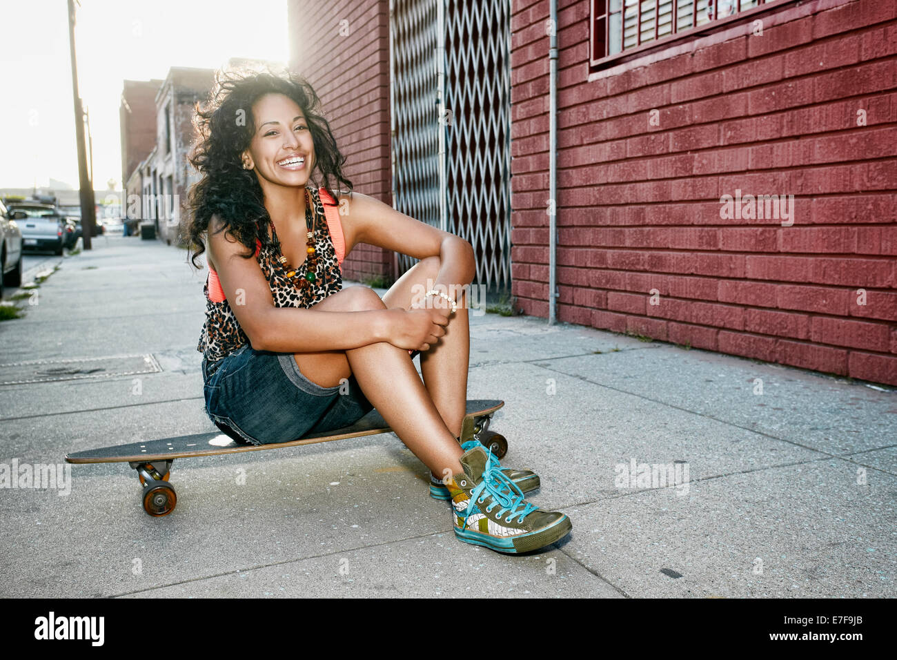 Hispanic woman sitting on skateboard on city street Stock Photo