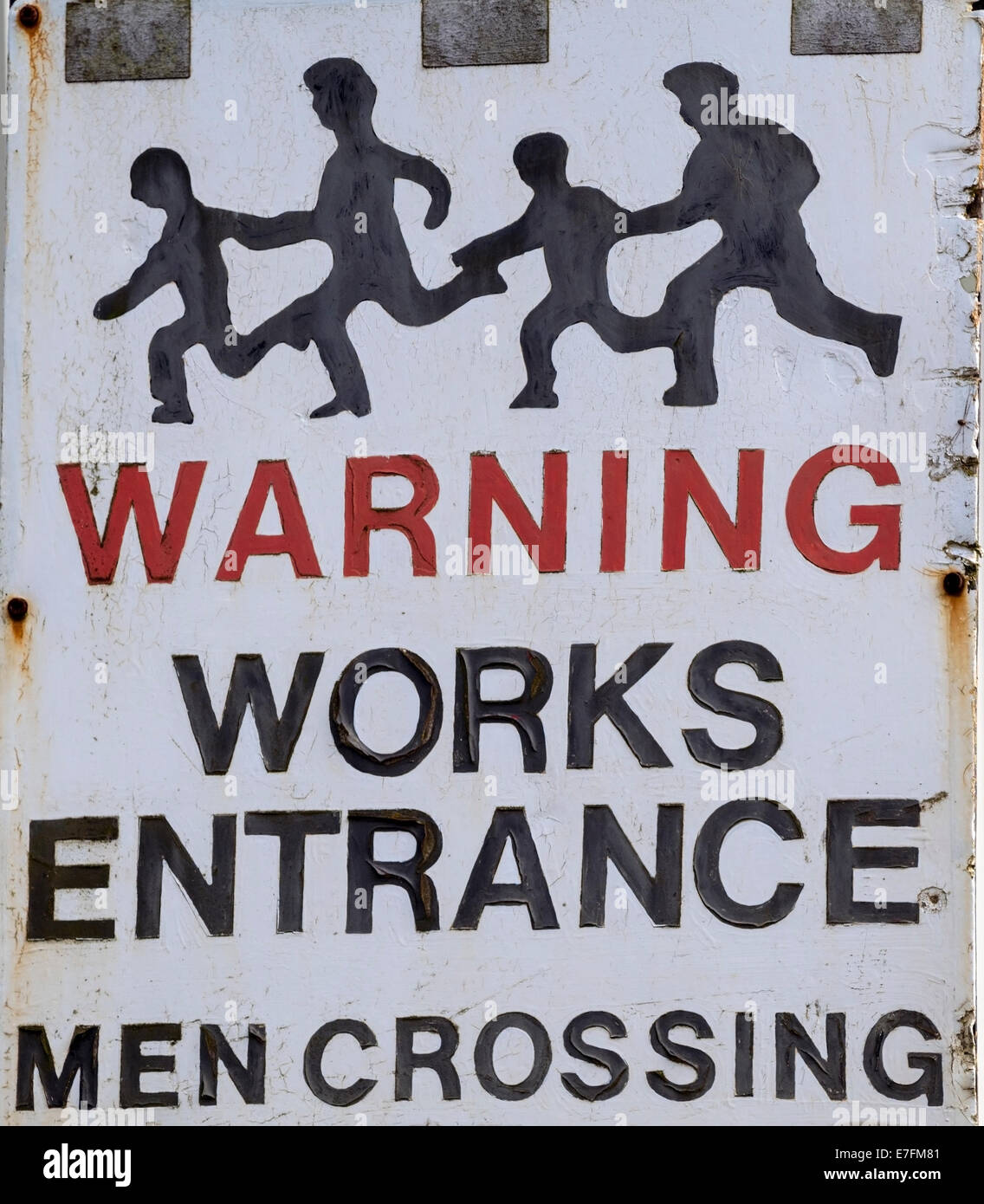 A hand painted road sign warning of workers crossing the road near a works entrance. Stock Photo