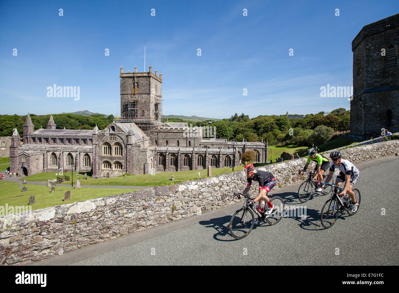 Three cyclists ride along a path by a stone church in Pembrokeshire, Wales Stock Photo