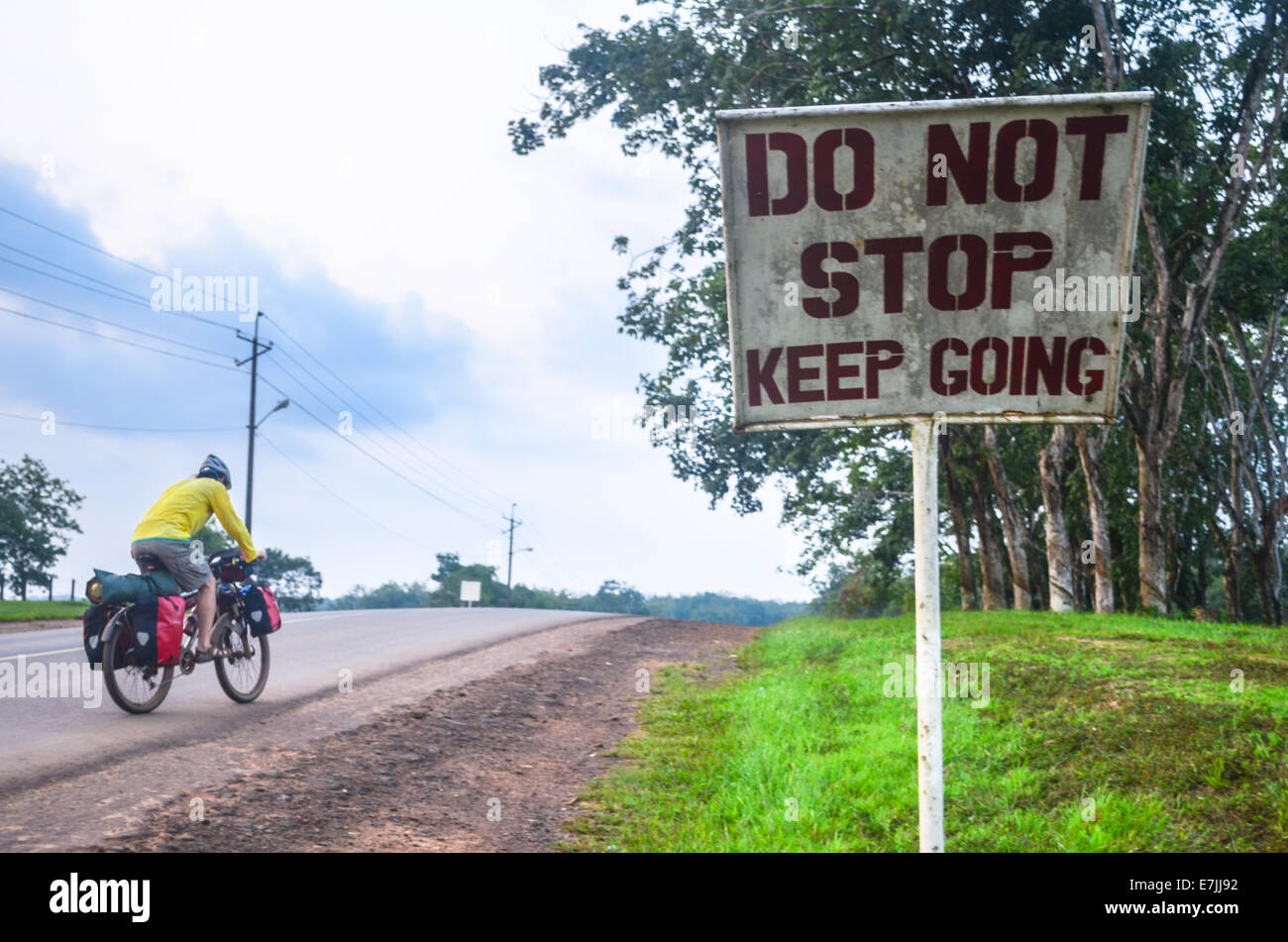 Touring cyclist cycling on a slope and passing a sign reading 'Do not stop keep going' near Monrovia, Liberia Stock Photo