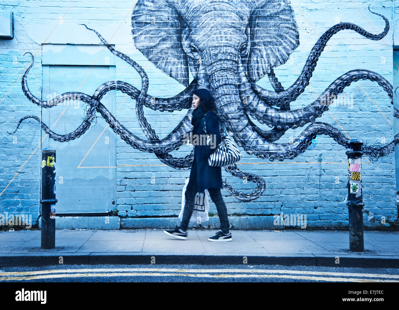 A girl walking on the street passing a graffiti with an elephant/octopus on a brick wall. Stock Photo