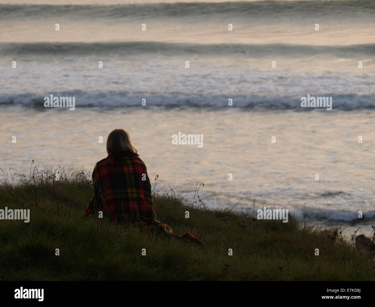 Woman wrapped in a blanket sat on the cliff top watching the sunsetting over the sea, Northcott, Bude, Cornwall, UK Stock Photo