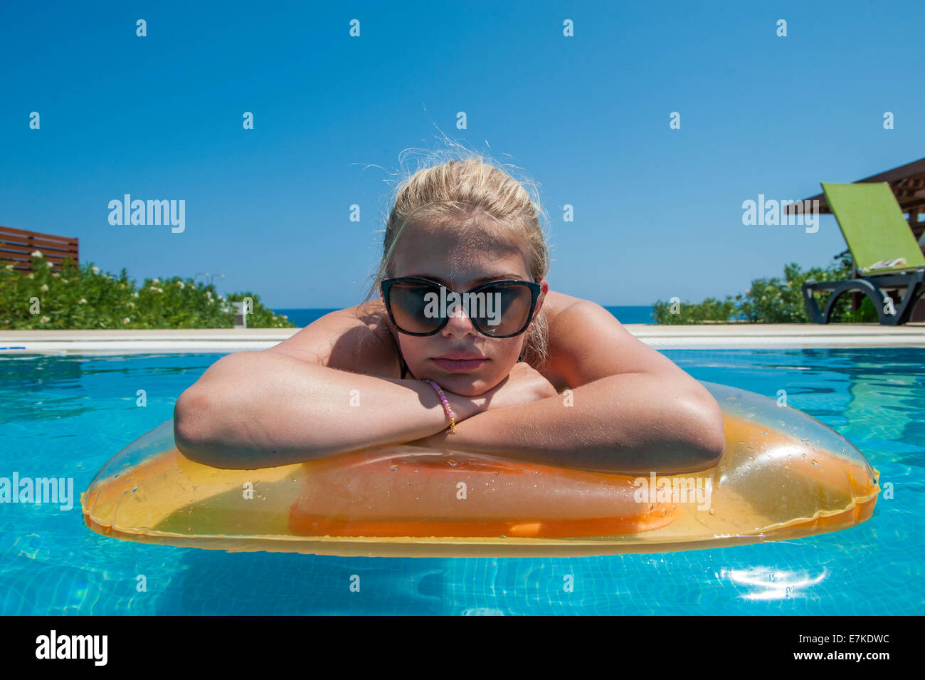 A blond girl in a pink bikini and sunglasses lies on an inflatable bed in a swimming pool in the hot sunshine Stock Photo