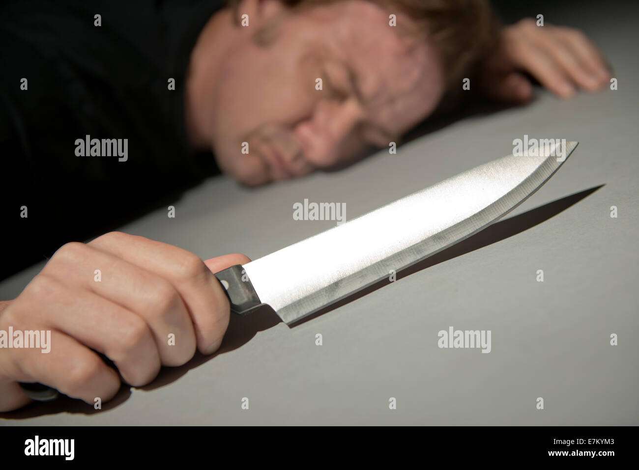 Angled view of a man, lying face down on the floor, holding a deadly kitchen knife. Stock Photo