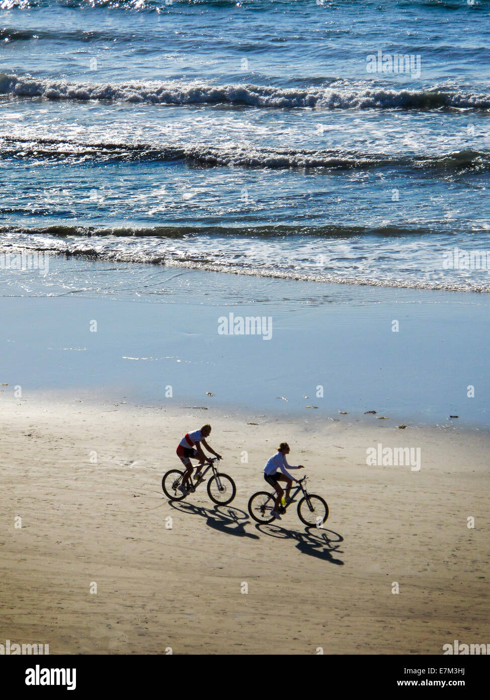 Throwing shadows in the afternoon sunlight, two bicyclists ride next to the Pacific Ocean surf in Laguna Beach, CA. Stock Photo