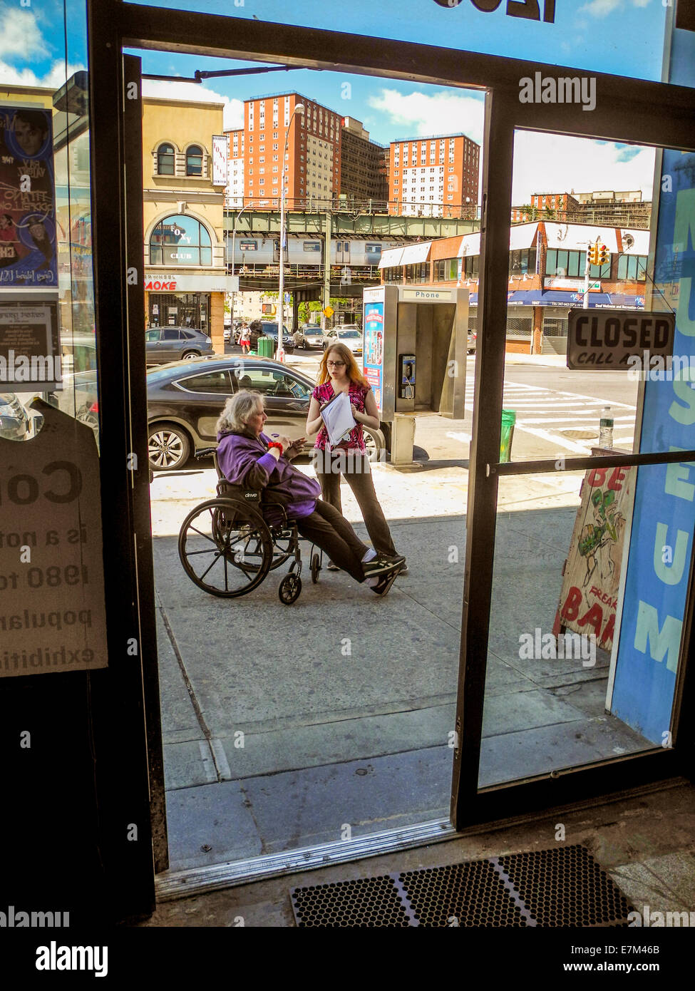 Seen through the door of her attraction, a handicapped concessionaire and her employee talk on the sidewalk in Coney Island, New York City. Note subway station and apartment houses at this famous amusement park. Stock Photo