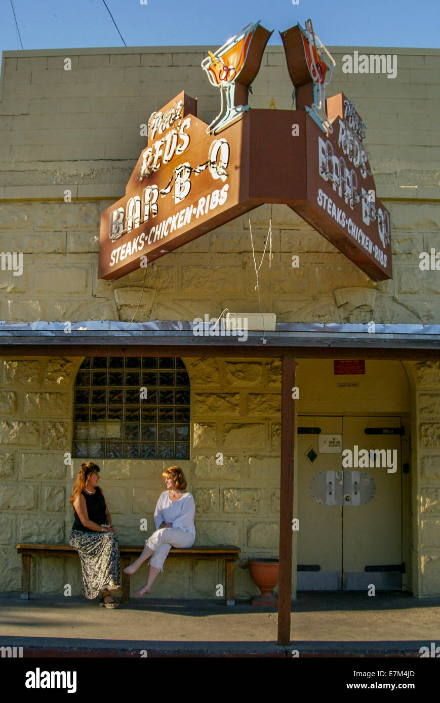 In late afternoon sun, two women talk while waiting for a bar-b-q restaurant to open in rural Diamond Springs, CA. Note cocktail sign. Stock Photo