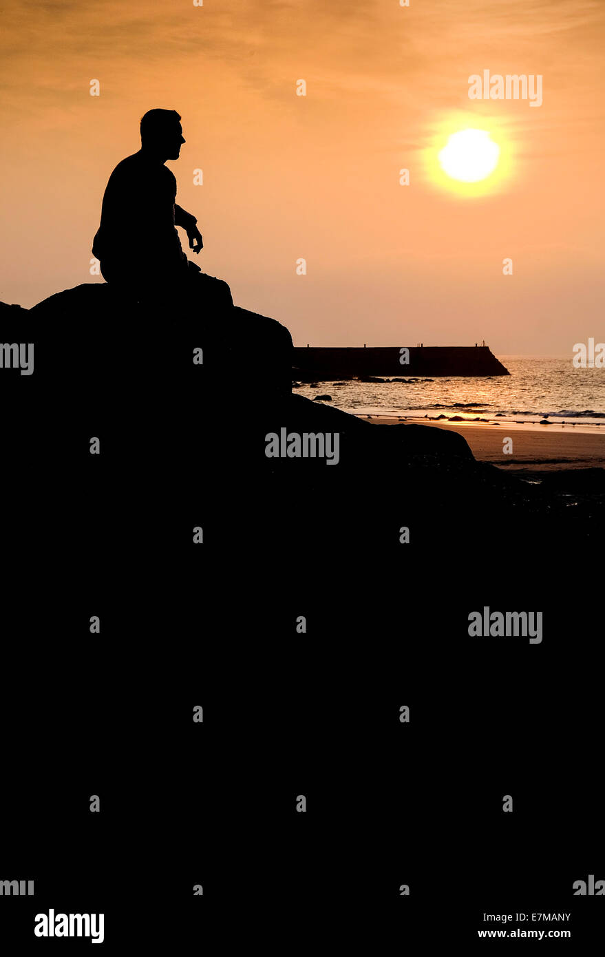 The silhouette of a man sitting on a rock at Sennen Cove as the sun sets. Stock Photo