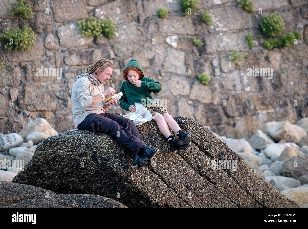 Holidaymakers eating fish and chips on rocks in Sennen Cove in Cornwall. Stock Photo