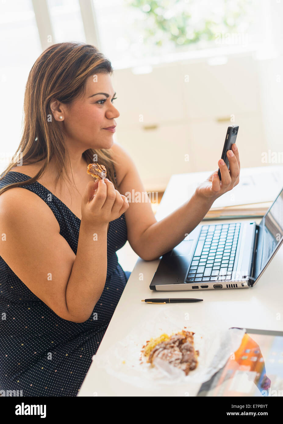 Woman eating croissant and using mobile phone in office Stock Photo