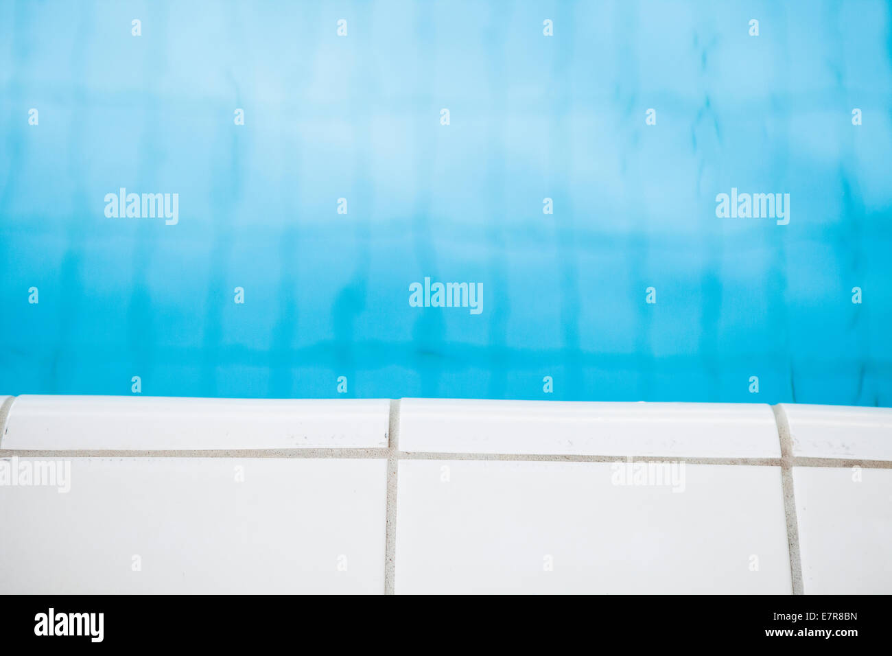 Closeup of white tiles at edge of swimming pool with blue water in background Stock Photo