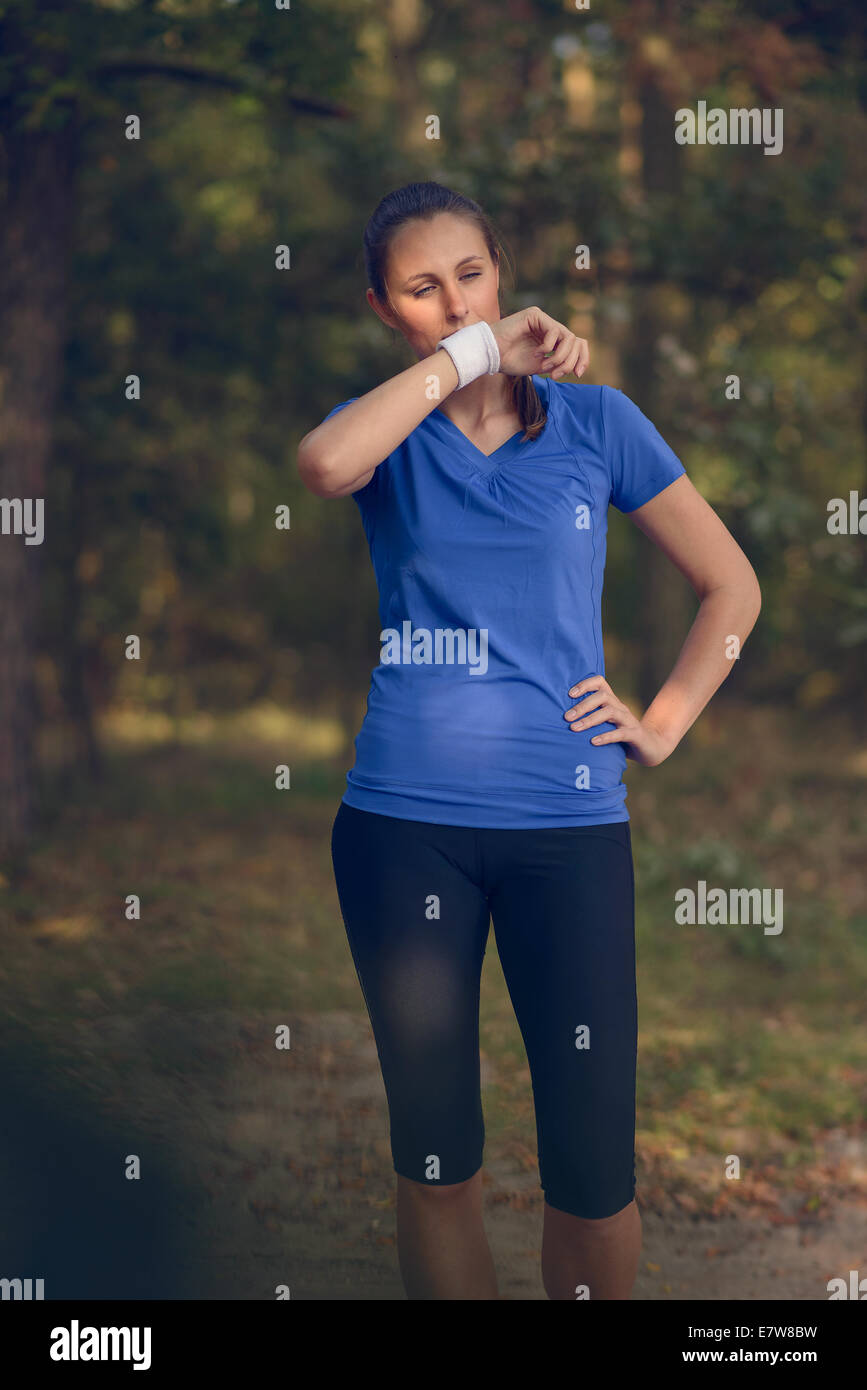 Woman athlete wiping sweat from her forehead onto her wristband as she pauses during her training exercises on a forest track Stock Photo