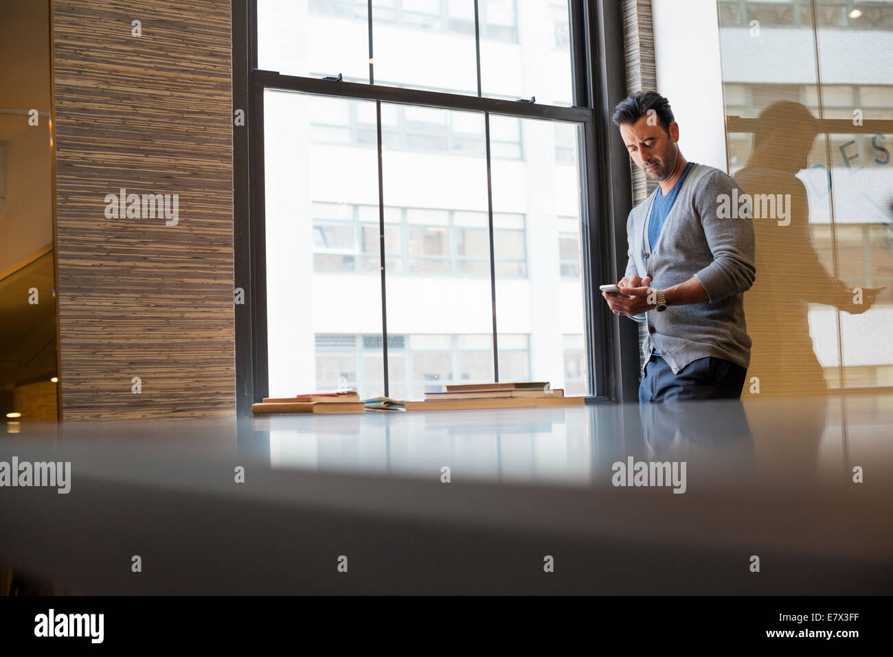 Office life. A man standing by a window in an office checking his smart phone. Stock Photo