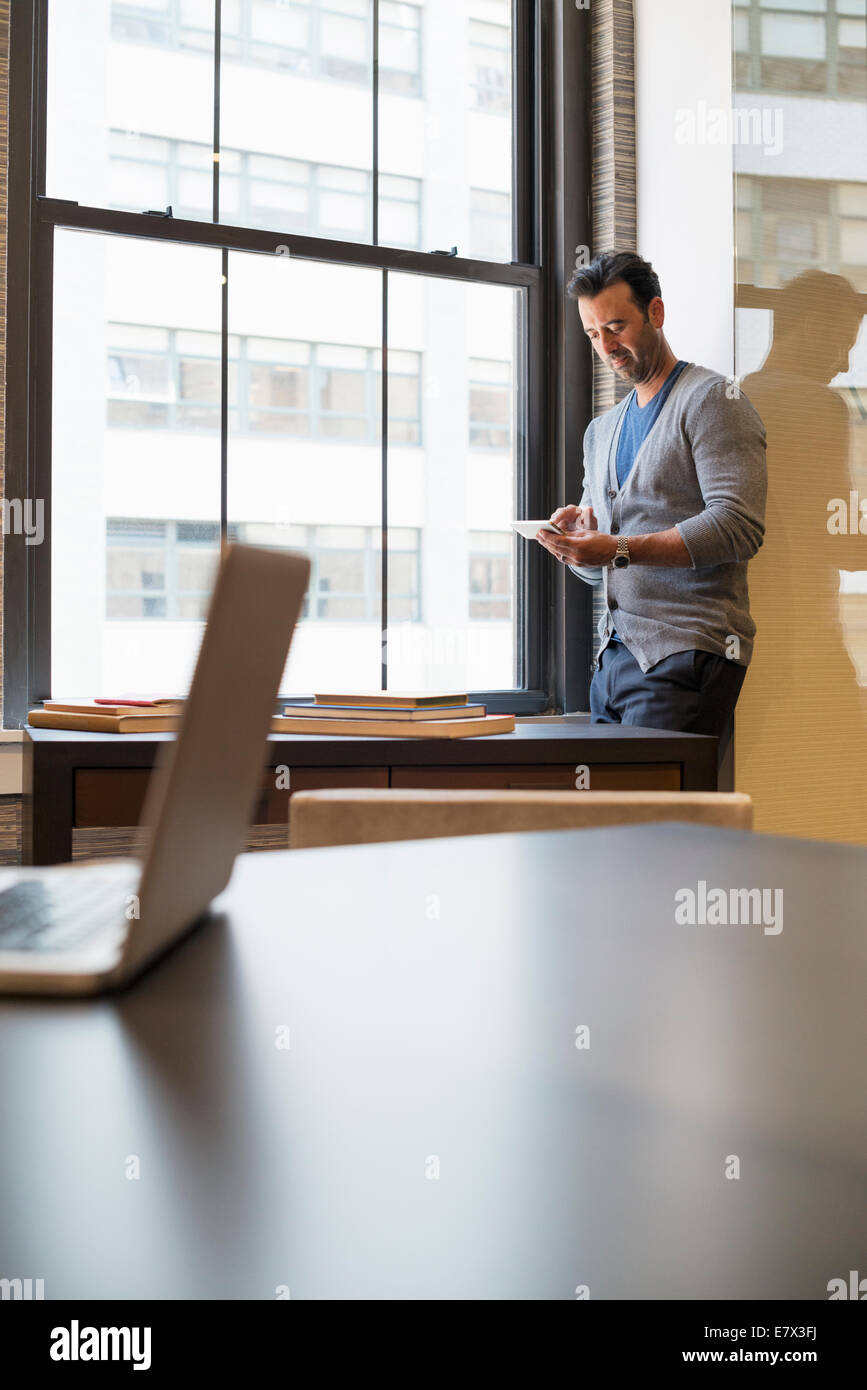 Office life. A man standing by a window in an office checking his smart phone. Stock Photo