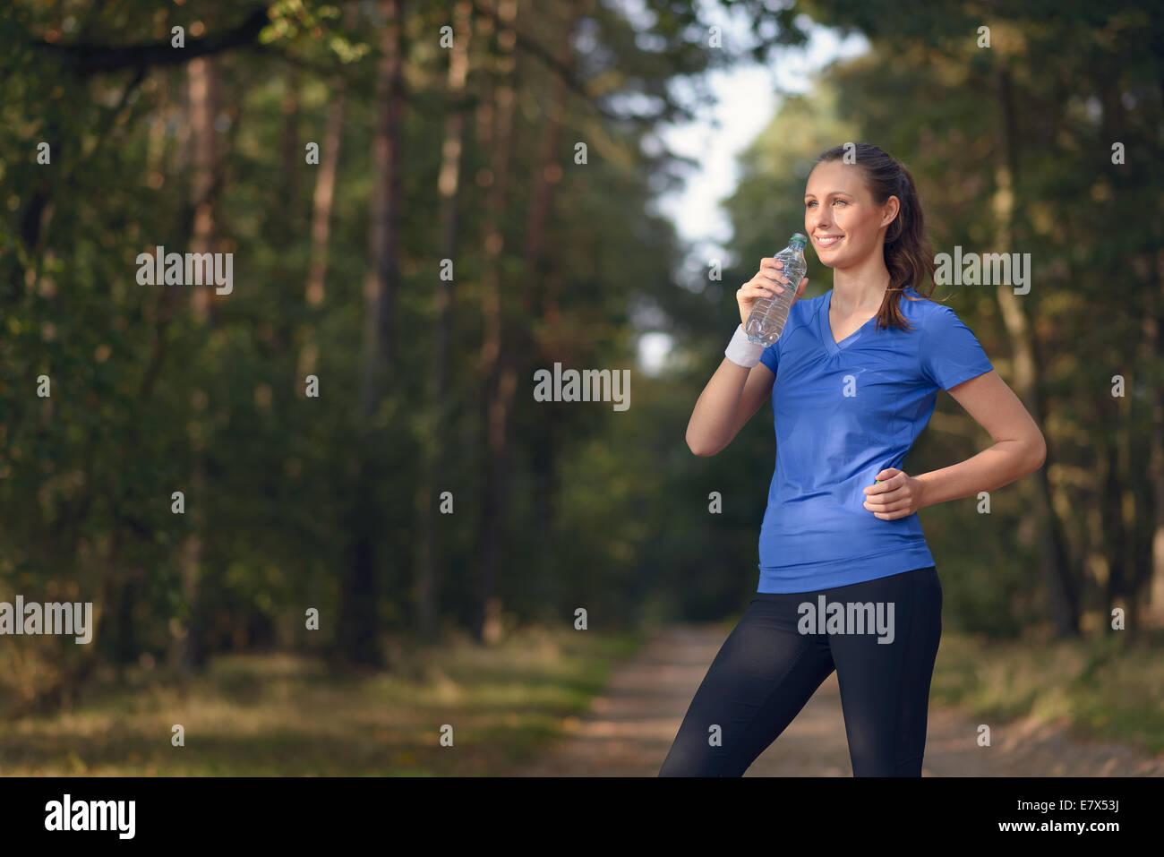 Fit slender young woman in sportswear drinking bottled water as she pauses on a forest track to re-hydrate during a training run Stock Photo