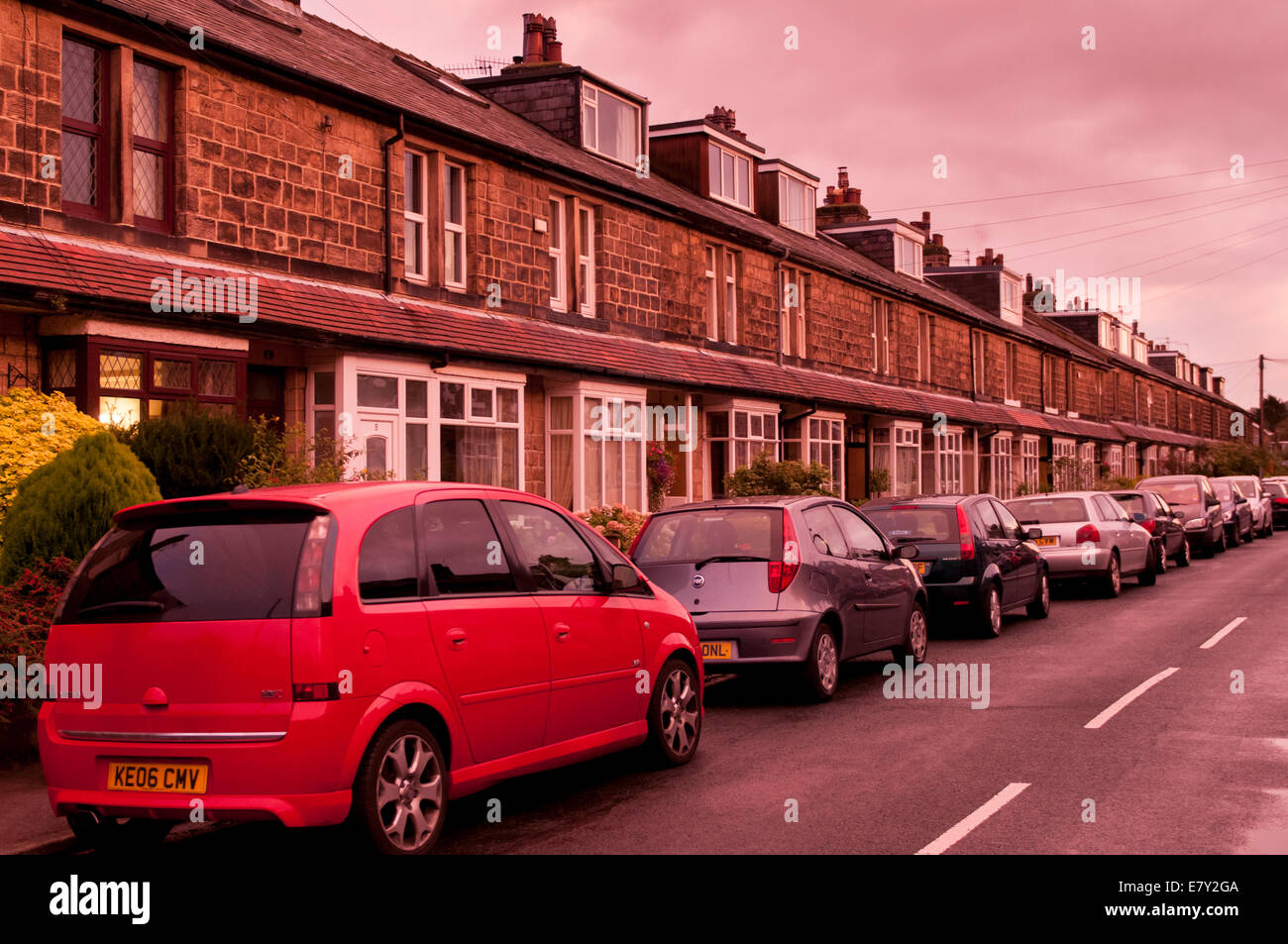 Continuous line of parked cars outside a long straight terrace of stone-built houses under a pink red sunset sky - Burley in Wharfedale, Yorkshire, UK Stock Photo