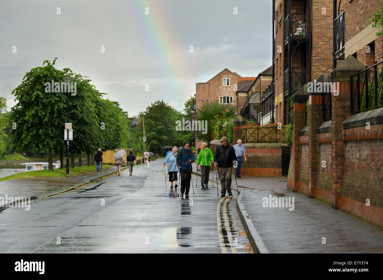 Rainbow high in grey cloudy sky after sudden downpour of rain soaked & wet people walking on riverside road - York, North Yorkshire, England, UK, Stock Photo