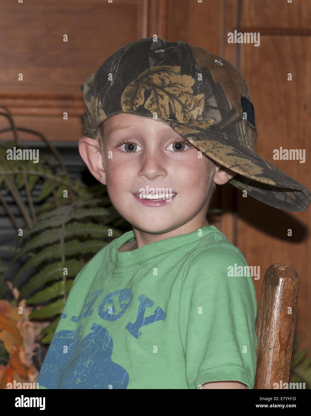 Boy child sitting in a chair inside looking sideways at the camera.  He has on a camo hat that is too big. Stock Photo
