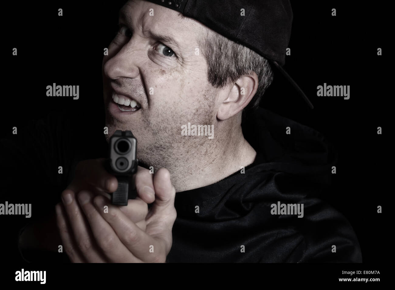 Closeup front view of mature man, looking forward and wearing baseball cap backwards, with gun in hand on dark background Stock Photo
