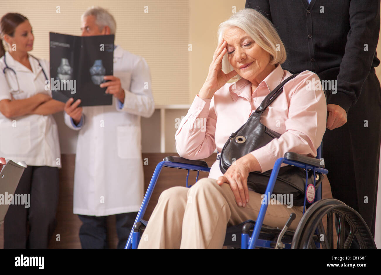 Mature woman on the wheelchair in hospital worried with her husband. Doctors analyzing scan in background. Stock Photo