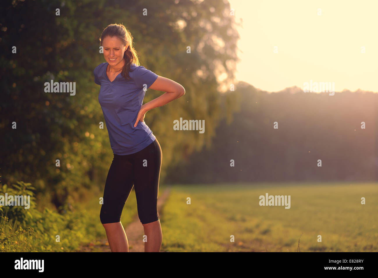 Woman athlete pausing to relieve her back pain holding her hand to her lower back with a grimace while out training Stock Photo