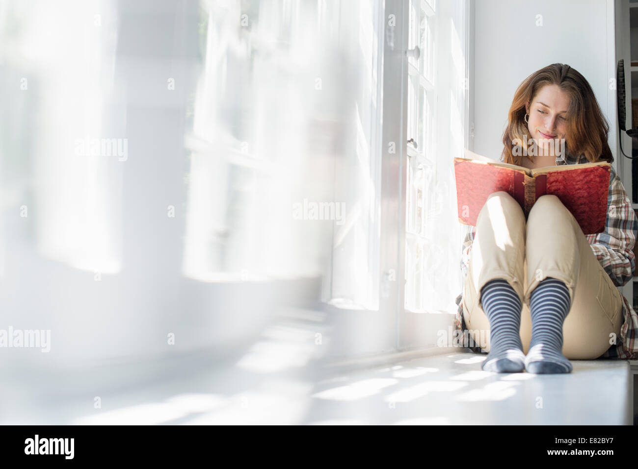 A woman seated by a window, reading with a book on her lap. Stock Photo