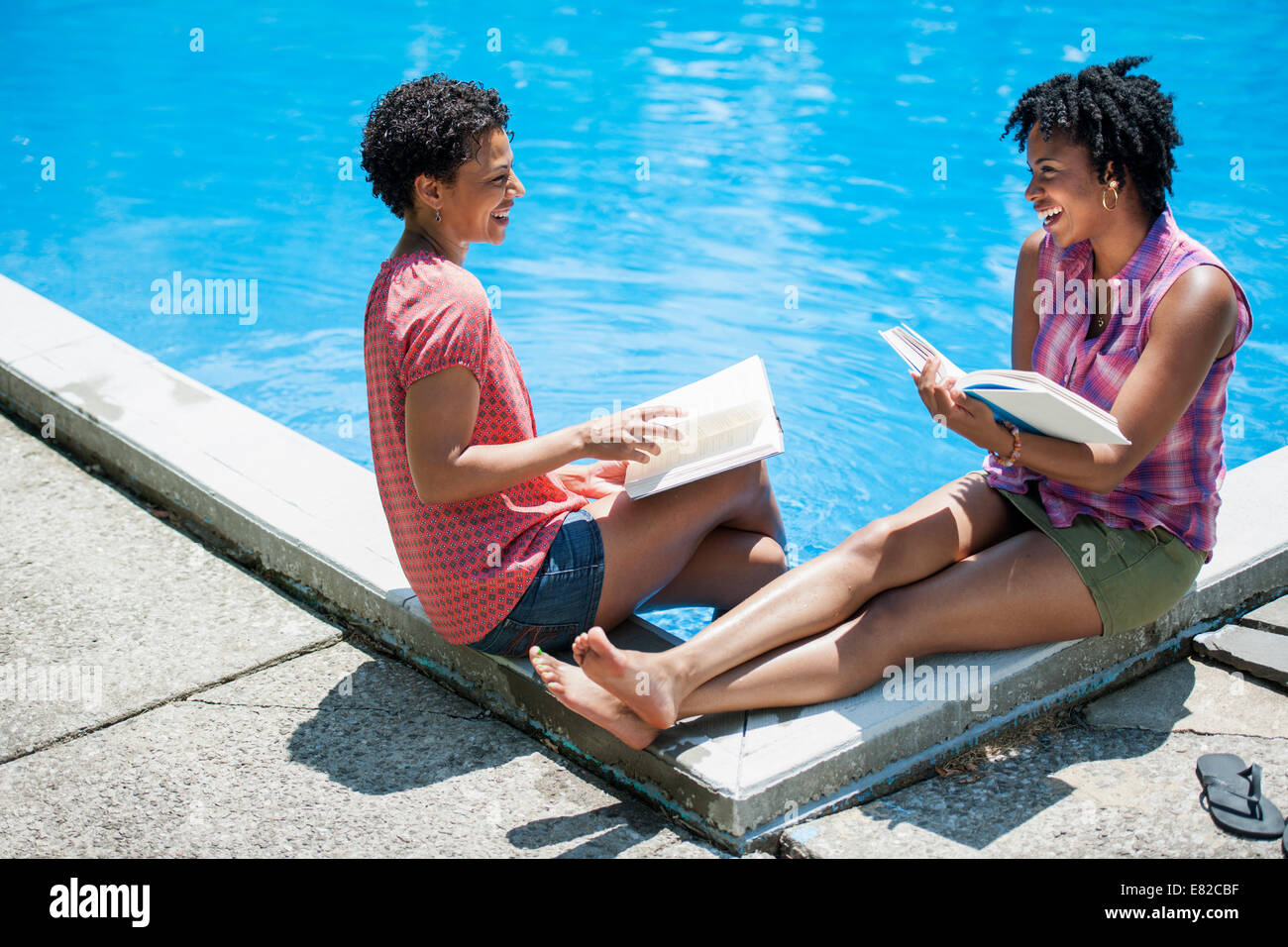 Two women sitting by the side of a swimming pool, reading books. Stock Photo