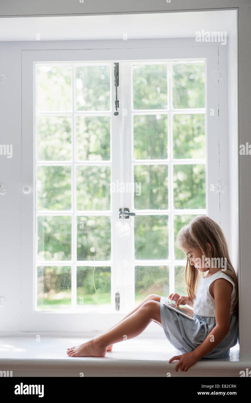 A young girl sitting at a window seat, using a digital tablet. Stock Photo
