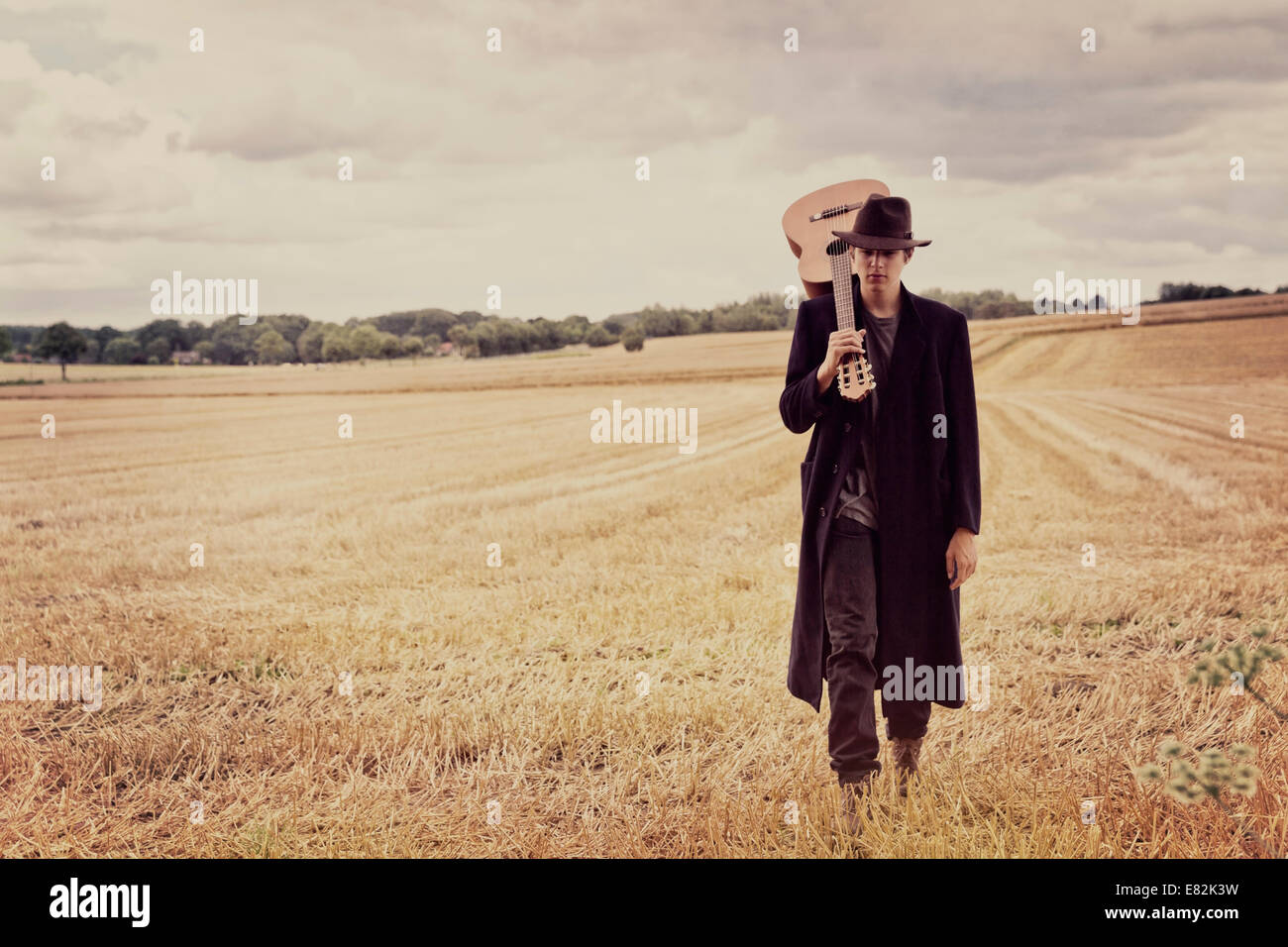 Teenage boy with hat and long coat walking on a grainfield with guitar Stock Photo