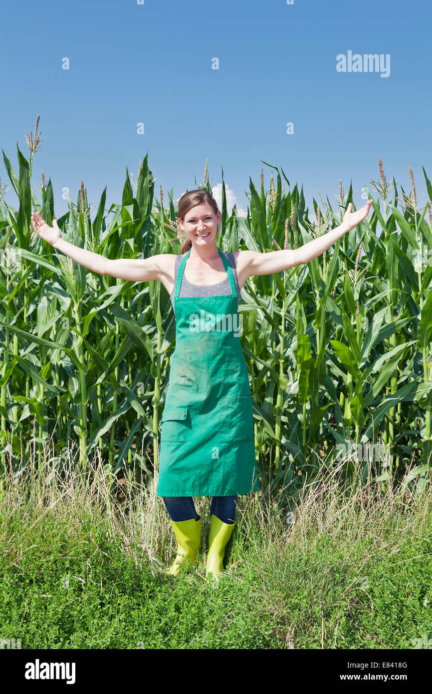 Young woman in work clothes standing with outstretched arms in front of a maize field, Baden-Württemberg, Germany Stock Photo