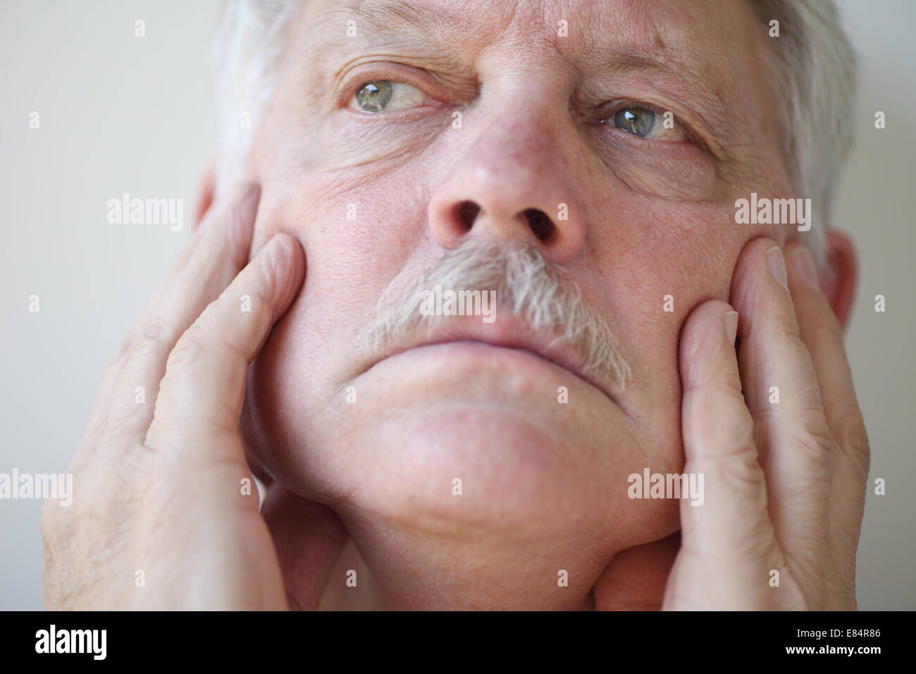 An older man has his hands on his cheeks and a thoughtful expression. Stock Photo